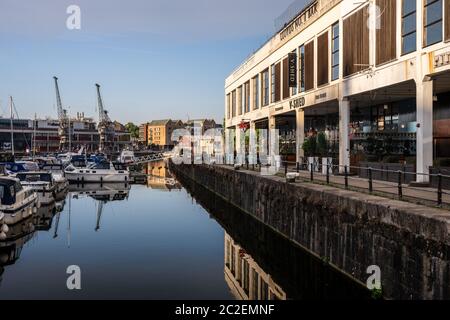 I joggers corrono sul Harbourside di Bristol accanto a V Shed, uno dei numerosi magazzini storici di capannoni di transito convertiti in bar e ristoranti. Foto Stock