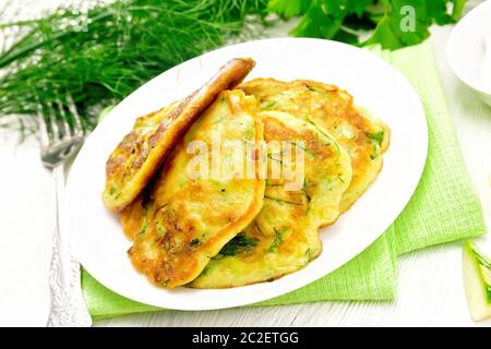 Frittelle di zucchini, aneto e prezzemolo nella piastra su un asciugamano, panna acida in un piattino sulla tavola di legno sfondo Foto Stock