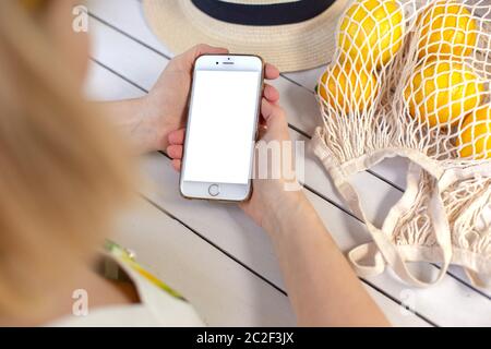 Mani di una ragazza che tiene uno smartphone bianco, su un tavolo di legno bianco, accanto ai limoni gialli in una borsa a maglia. Vista dall'alto. Bock up Foto Stock