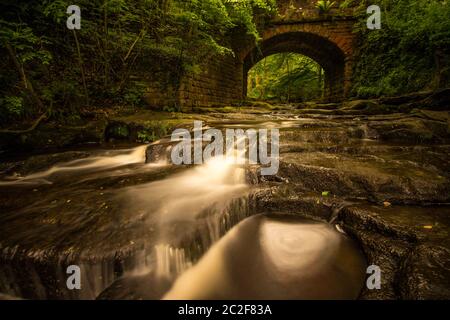 Piccolo ponte di mattoni rossi sul torrente in cascata foss. Flusso dolce con piscine e increspature Foto Stock