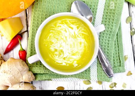 Zuppa di purea di zucca con la crema in una ciotola bianco su un asciugamano da cucina con un cucchiaio, zucca, baccelli di caldo rosso pepe, aglio, radice di zenzero su una tavola di legno Foto Stock