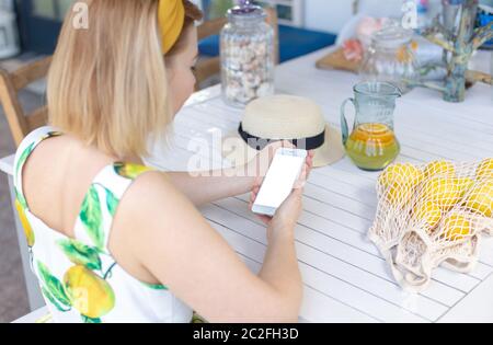 Una ragazza bionda in una semina estiva si siede ad un tavolo bianco, tiene uno smartphone bianco nelle sue mani, accanto ad esso è una brocca di limonata e limoni gialli dentro Foto Stock