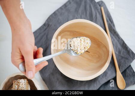 Donna che tiene un cucchiaio di avena sopra una ciotola di legno su un tovagliolo su una superficie bianca Foto Stock