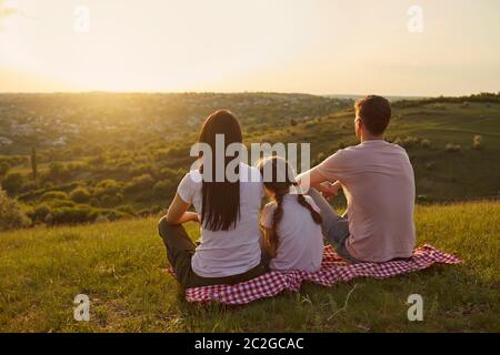 Vista posteriore della famiglia giovane con bambino seduto su coperta da picnic e guardando il tramonto bello in montagna, spazio copia Foto Stock