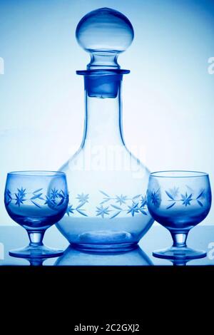 Decanter di alcool di cristallo vuoto e due bicchieri per la vodka in piedi su un tavolo di vetro. Vista laterale verticale, tono freddo. Foto Stock