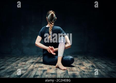 Vista posteriore di giovane donna che fa yoga asana mezzo signore dei pesci posa in camera oscura. Foto Stock