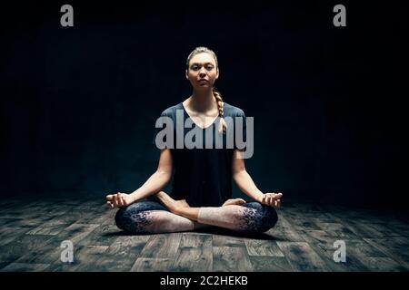 Giovane donna attraente che pratica yoga seduta in posa di loto e meditando in camera oscura Foto Stock