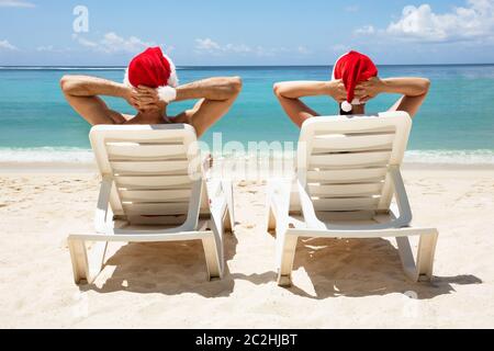 Vista posteriore di un paio di indossare cappelli di Babbo Natale rilassante su bianco Sedie a sdraio sulla spiaggia Foto Stock