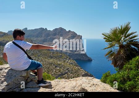 Ragazzo latino, felice, con i capelli scuri, in posa, ritratto, in una giornata fuori con la sua famiglia in primavera, seduto sulla scogliera di Cap de Formentor, indicando Foto Stock