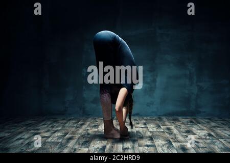 Giovane donna che fa yoga in piedi in avanti piega posa in camera oscura Foto Stock