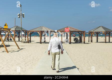 Un uomo anziano in un cappello piatto, camicia bianca e pantaloni che cammina su una spiaggia a Tel Aviv, Israele Foto Stock