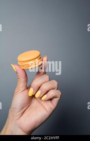 Primo piano di una mano di donna che tiene su una pasta di macaron francese arancione su uno sfondo scuro. Foto Stock