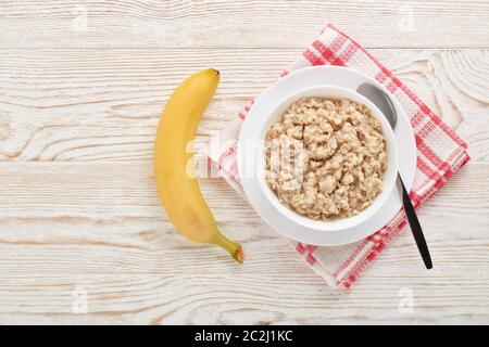 Porridge di farina d'avena in ciotola bianca e banana su sfondo di legno chiaro. Vista dall'alto con spazio per la copia. Concetto di stile di vita sano, cibo sano, dieta, Foto Stock