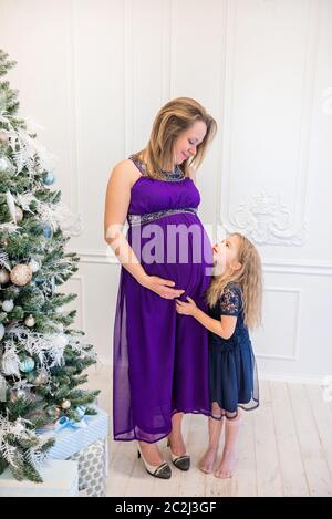 Bella donna incinta in ultra abito viola in piedi con la figlia sul divano vicino a albero di natale Foto Stock