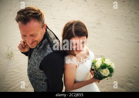Vista in primo piano su sposa e sposo sorridente e in piedi sulla spiaggia di sabbia e guardando la stessa direzione. Sposi alla luce della sera. Giorno di nozze Foto Stock