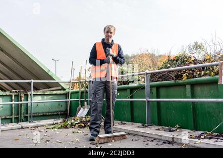 Uomo che spazzano il pavimento del centro di riciclaggio dopo aver portato i rifiuti verdi nel contenitore Foto Stock