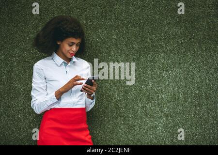 Una donna afro-americana affascinante con un cellulare è in piedi davanti ad un muro verde di erba artificiale; giovane donna d'affari biraciale Foto Stock