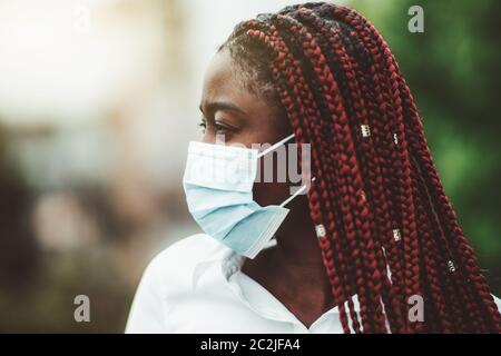 Primo piano ritratto di una giovane donna africana con trecce di castagne e in una maschera protettiva contro il virus sopra il viso; maschera nera all'aperto - protezione Foto Stock