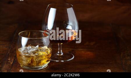 Whisky on the rocks e cognac in un snifter in piedi su una tavola di legno banco bar di notte con spazio di copia Foto Stock