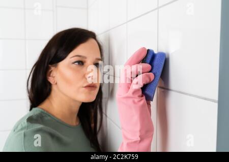 Giovane donna con occhiali protettivi Pulizia della piastrella bianca della parete utilizzando una spugna Foto Stock