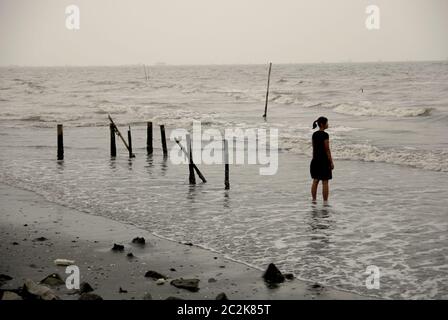 Donna che guarda verso la Baia di Giacarta mentre si trova sulla spiaggia sabbiosa durante una giornata piovosa a Marunda, Jakarta Nord, Giacarta, Indonesia. Foto Stock