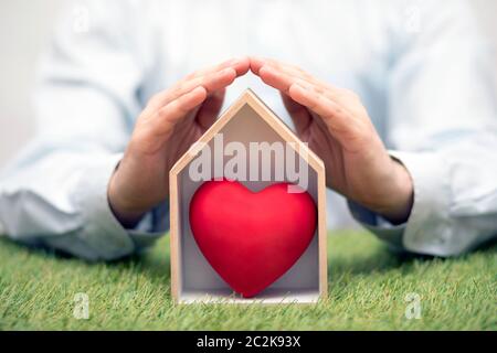 Casa in legno con cuore rosso su erba verde protetto dalle mani Foto Stock