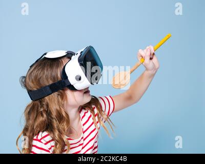 sorridente positivo ragazza che indossa occhiali di realtà virtuale occhiali di protezione cuffie sta cantando con cucchiaio da cucina, nuova generazione, concetto di fronte a sfondo blu Foto Stock