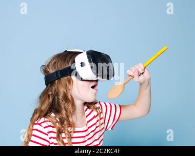 sorridente positivo ragazza che indossa occhiali di realtà virtuale occhiali di protezione cuffie sta cantando con cucchiaio da cucina, nuova generazione, concetto di fronte a sfondo blu Foto Stock