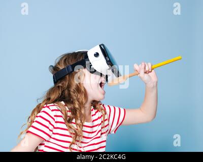 sorridente positivo ragazza che indossa occhiali di realtà virtuale occhiali di protezione cuffie sta cantando con cucchiaio da cucina, nuova generazione, concetto di fronte a sfondo blu Foto Stock