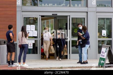 Baltimora, Maryland, Stati Uniti. 17 Giugno 2020. Due clienti femminili con maschere facciali che trasportano sacchetti di carta di generi alimentari emergono dalla sede di Baltimore Harbor East Whole Foods Market al 711 South Central Avenue il giorno di apertura, mentre altri clienti attendono il permesso dal personale che controlla le porte per entrare; Un segno "abbiamo spostato" con il logo accanto a loro. La precedente sede di questa filiale della catena di supermercati multinazionale di proprietà di Amazon, chiusa alcuni giorni prima, era situata nella vicina Fleet Street. Kay Howell/Alamy Live News Foto Stock