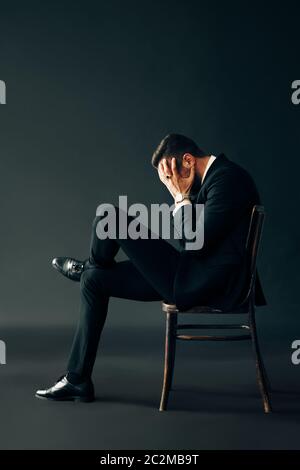 Triste uomo infelice in abito nero seduto sulla sedia e chiuso il viso da due mani Foto Stock
