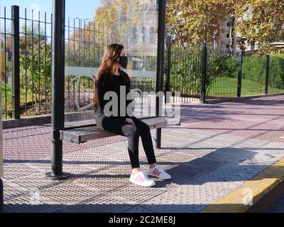 Millennial Brunette ragazza in cotone maschera seduta ad una fermata vuota del bus, in attesa da sola a causa del virus Corona COVID 19 Pandemic Lockdown. In maschera medica Foto Stock