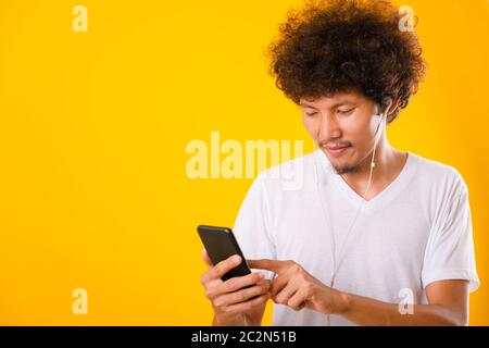 Felice asiatico bello uomo con capelli ricci sorridente godendo di ascoltare la musica sulle cuffie utilizzando uno smartphone cellulare isolato su sfondo giallo Foto Stock