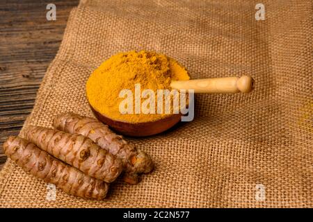 Radici turmeriche fresche intere e polvere di curcuma essiccata in una ciotola di legno con un cucchiaio di legno davanti ad uno sfondo con un tessuto di iuta. Foto Stock