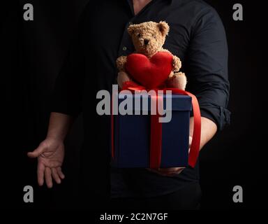 Uomo adulto in una camicia nera contiene un quadrato blu box legate con un nastro rosso e si siede sulla parte superiore di un marrone orsacchiotto di peluche con un cuore, concetto di congratulatio Foto Stock