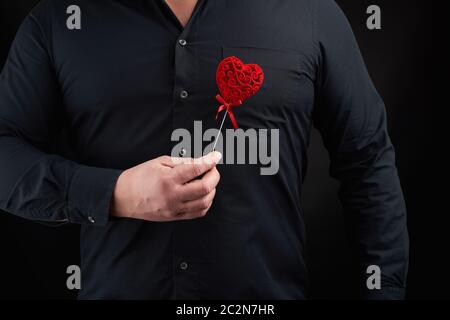 Adulto Uomo sorge su uno sfondo scuro che indossa una camicia nera e tenendo un rosso cuore intagliato nei pressi del suo petto nella sua mano destra, il concetto di amore Foto Stock