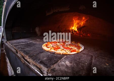 Professional forno a pizza. Un buon fuoco arde nel forno. Pizza con prosciutto e pomodori al forno. Per la cottura della pizza in pizzeria. Foto Stock