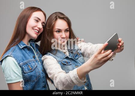 Due ragazze sorridenti in giacche denum rende selfie sulla fotocamera del telefono in studio. Amicizia femminile. Tempo libero di felice ragazze Foto Stock