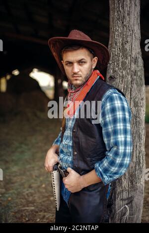 Brutale cowboy in jeans e giacca di pelle, texas ranch sullo sfondo, occidentale. Vintage persona di sesso maschile con il revolver wild west lifestyle Foto Stock