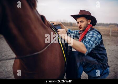 Brutale cowboy in jeans e giacca di pelle si arrampica a cavallo su ranch del Texas occidentale. Vintage persona di sesso maschile con cavallo selvaggio west Foto Stock