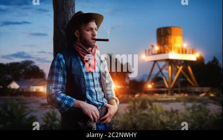 Brutale cowboy fuma un sigaro sul tramonto, relax serale sul ranch del Texas occidentale. Vintage persona di sesso maschile con pistola sulla fattoria, wild west cultura Foto Stock