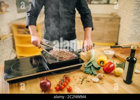 Uomo che cucina carne in una padella in cucina. Uomo che prepara maiale bollito su stufa elettrica della tavola Foto Stock