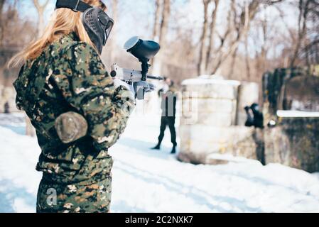 Paintball femmina di tiro del giocatore al nemico, vista posteriore, foresta invernale battaglia. Sport estremo gioco, i giocatori in maschere di protezione e mimetizzazione giace sul Foto Stock