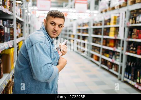 L uomo si nasconde una bottiglia di alcool sotto la sua camicia nel supermercato. Ladro maschio in negozio, marito conducendo una operazione segreta, shoplifling Foto Stock