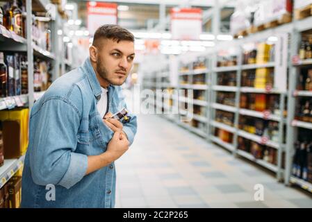 L uomo si nasconde una bottiglia di alcool sotto la sua camicia nel supermercato. Ladro maschio in negozio, marito conducendo una operazione segreta, famiglia shopping Foto Stock