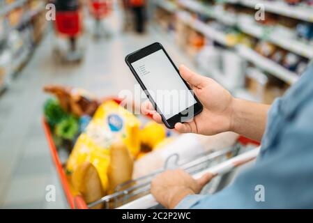 L'uomo con il carrello effettua un acquisto dall'elenco sul telefono nel supermercato. Maschio di cliente in negozio, non dimenticate di comprare nozione Foto Stock