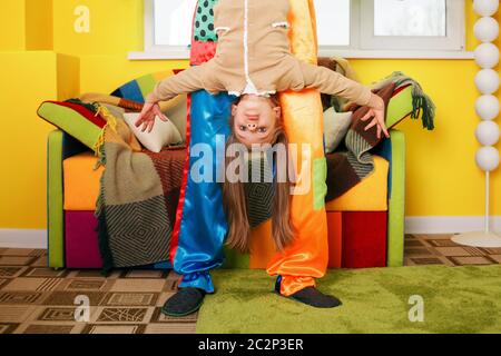 Il pagliaccio divertente tiene la testa della bambina sui tacchi. Divano colorato sullo sfondo. Foto Stock