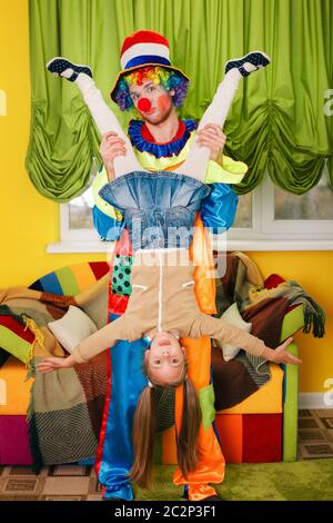 Il pagliaccio divertente tiene la testa della bambina sui tacchi. Divano colorato sullo sfondo. Foto Stock