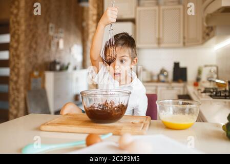 Bambino giovane che mescola il cioccolato fuso in una ciotola. Cucina carina ragazzo in cucina. Happy child prepara il dolce al banco Foto Stock
