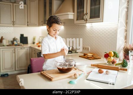 Ragazzo giovane che mescola il cioccolato fuso in una ciotola. Carino bambino maschile cucina in cucina. Happy child prepara il dolce al banco Foto Stock